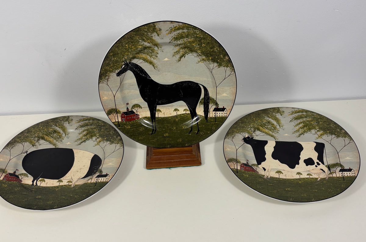 Three decorative ceramic plates with farm animal illustrations, including a central round plate featuring a black horse and two oval plates with a black-and-white horse and a black-and-white cow, displayed on a white surface against a light gray wall.