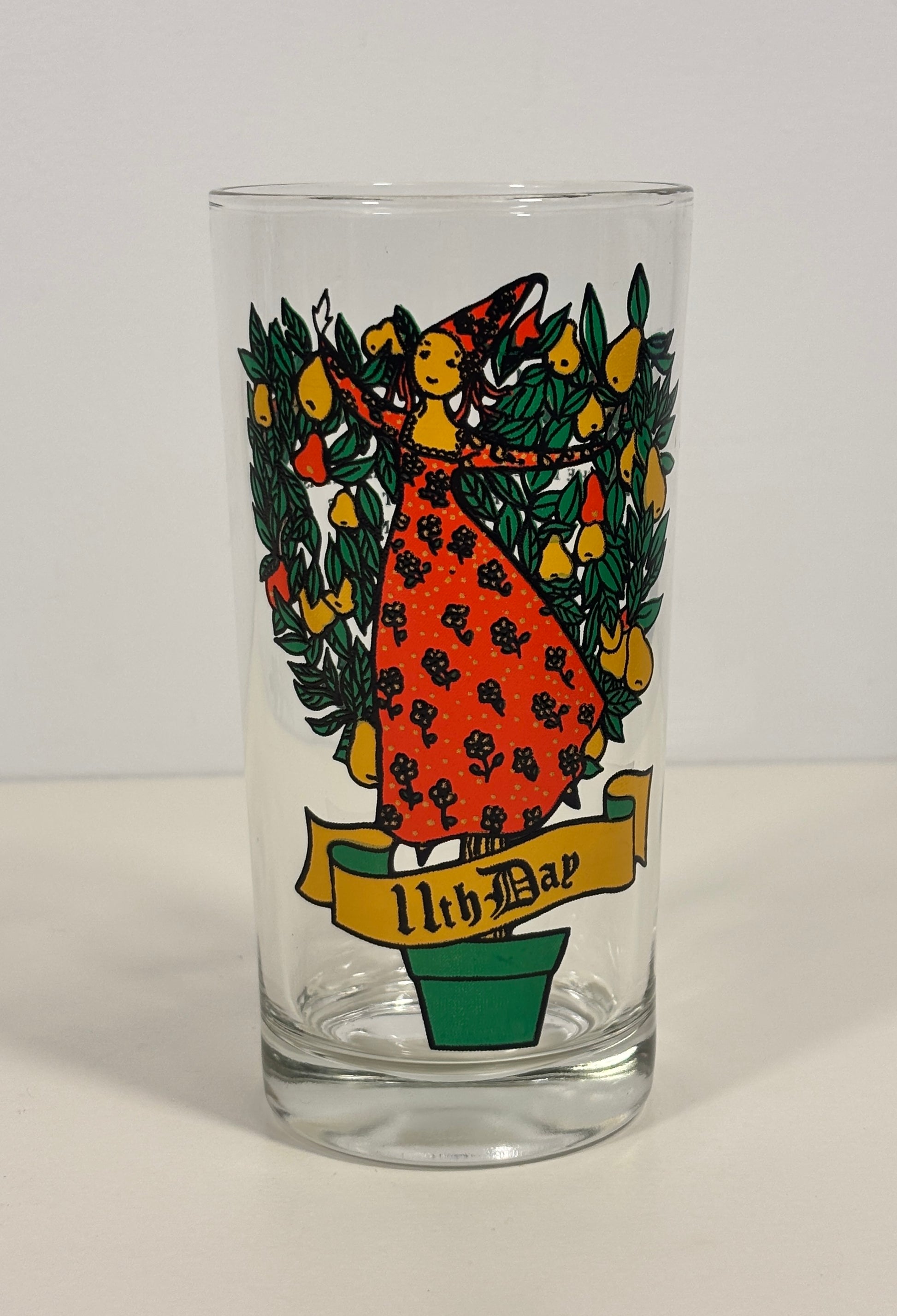 A clear glass highball tumbler featuring a colorful printed illustration of a woman in a red dress surrounded by green leaves and yellow fruit, with a yellow banner reading 'The 11th Bar' at the bottom.