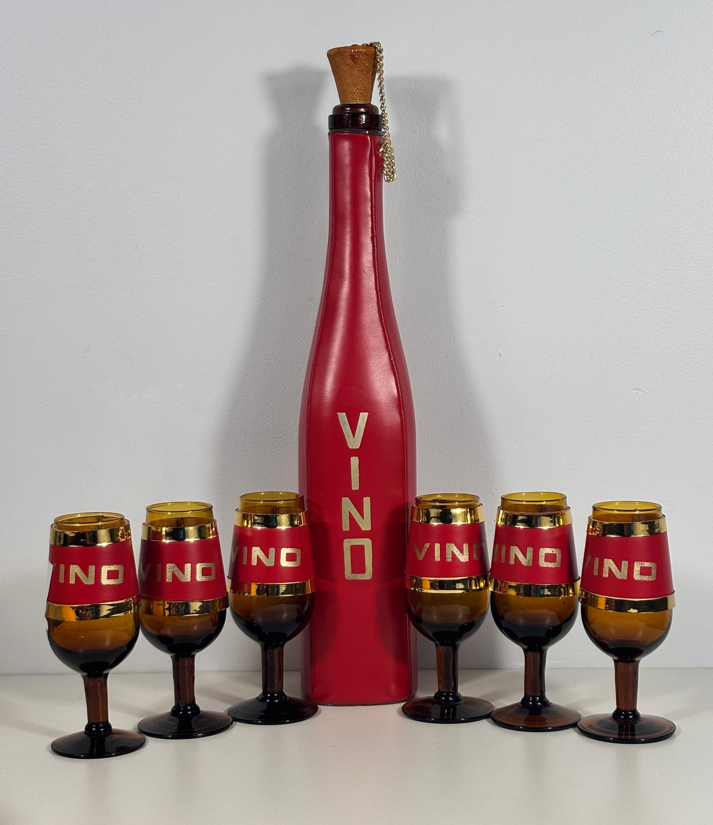 A vintage-style red wine bottle centerpiece with six matching amber and red wine glasses arranged in front on a white surface against a light gray background.