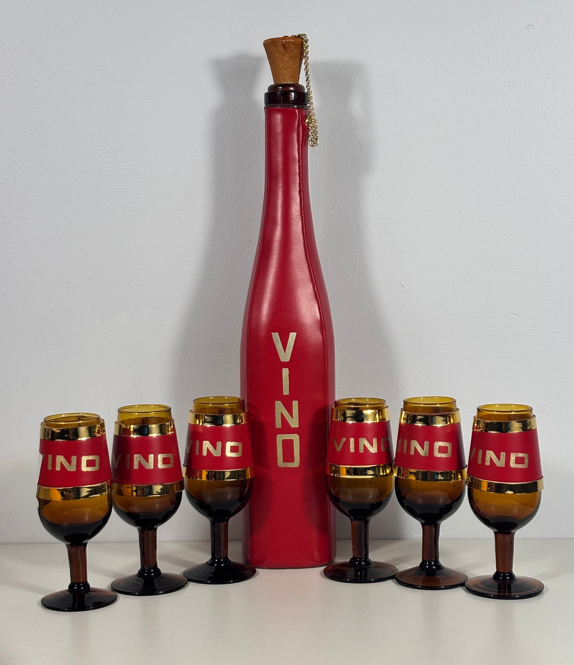A vintage-style red wine bottle centerpiece with six matching amber and red wine glasses arranged in front on a white surface against a light gray background.