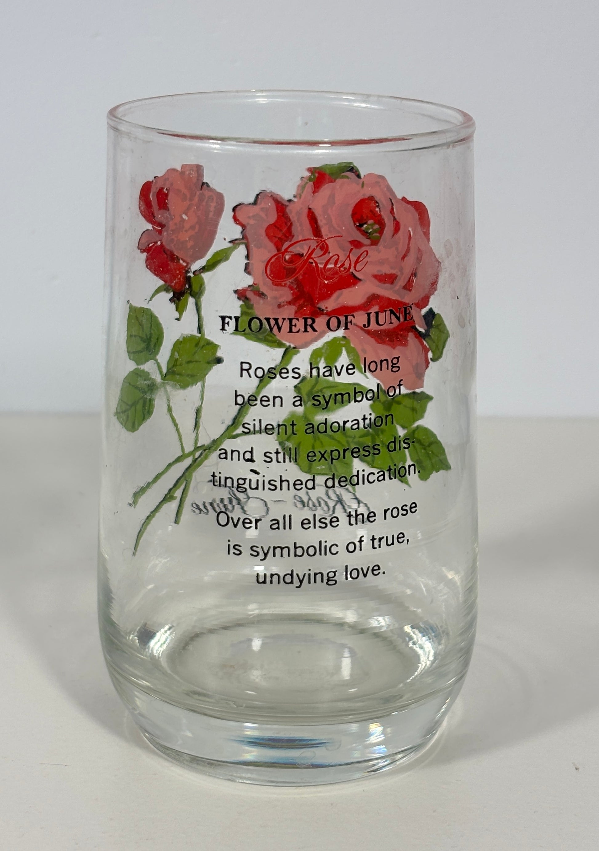 A clear glass tumbler featuring a vintage-style illustration of red and pink roses with green leaves, accompanied by the text 'FLOWER OF JUNE' and a poem about roses, set against a plain light background.