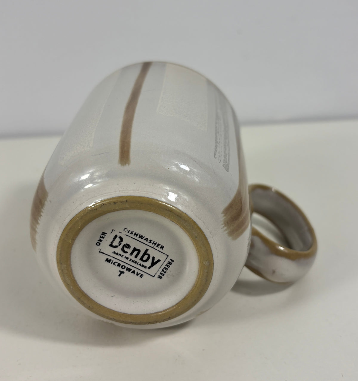A close-up shot of the bottom of a white ceramic mug with a brown handle and brown stripe details, showing the printed brand 'Denby' on the base.