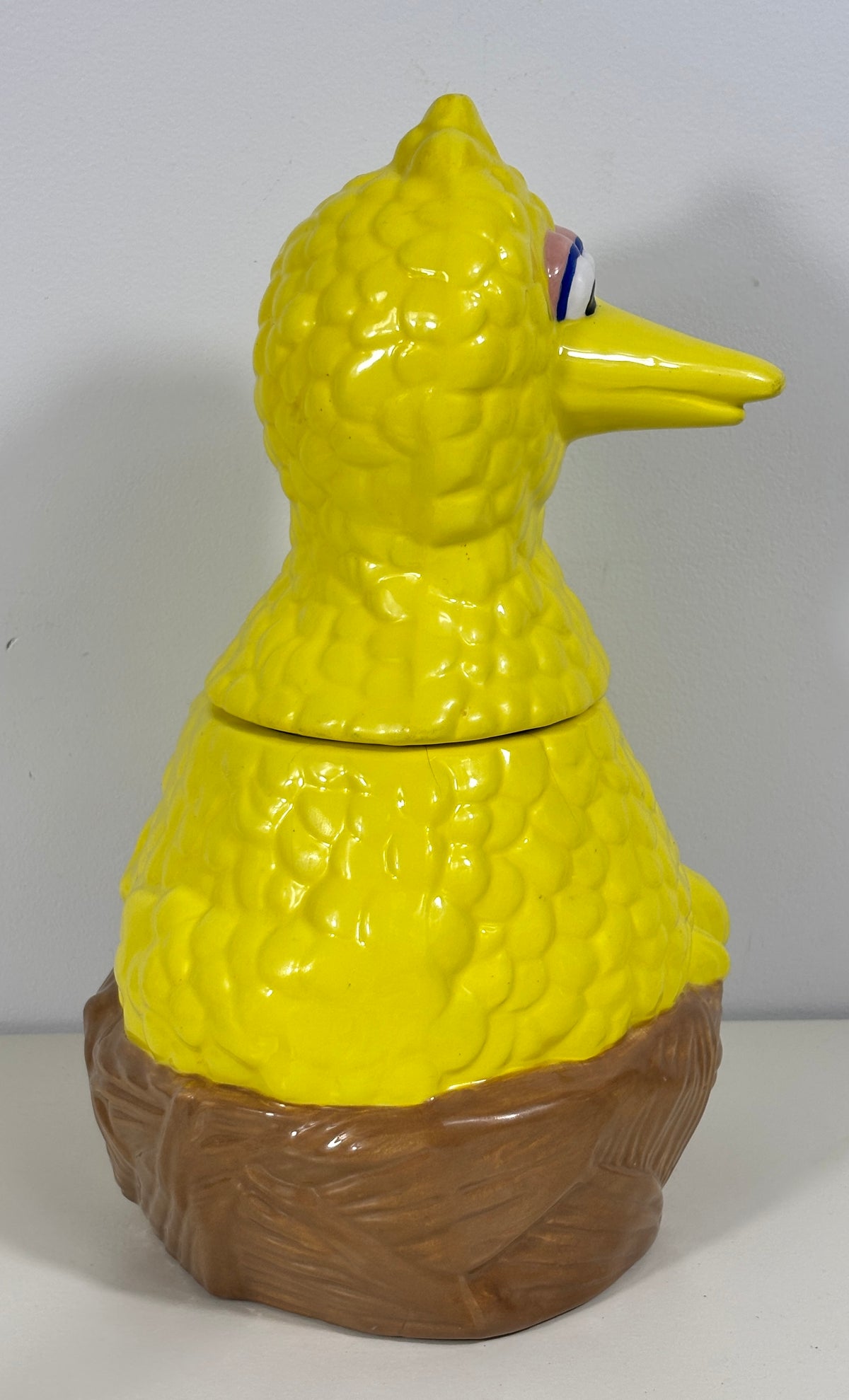 A yellow bird-shaped ceramic container with a textured, bumpy surface, featuring a brown textured base resembling a nest or tree trunk. The bird has a yellow beak and a small blue and red accent near its eye.