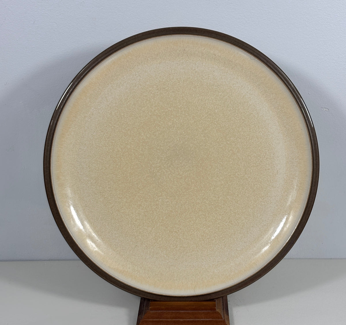 A round ceramic plate with a speckled cream-beige glaze and a contrasting dark brown rim, displayed on a wooden stand against a light gray background.