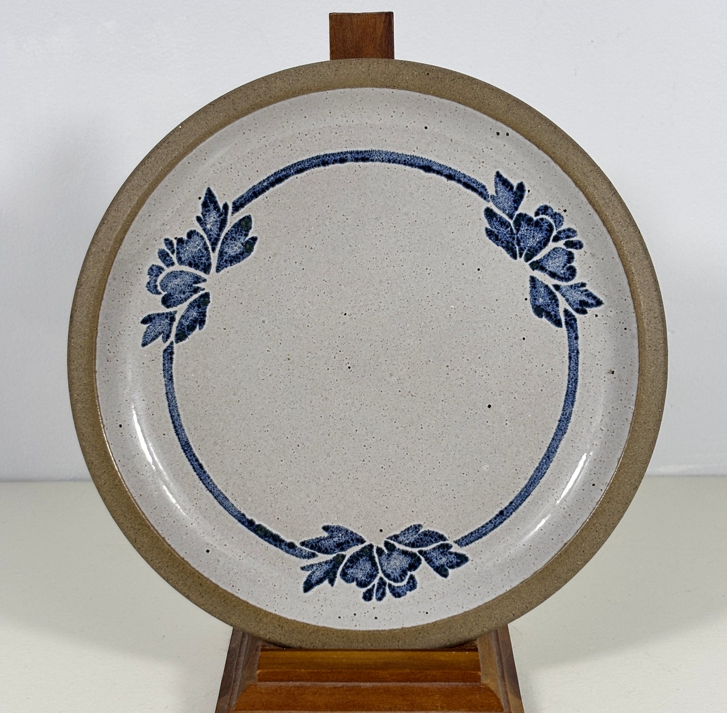 A round ceramic plate with a speckled off-white glaze, featuring a blue floral wreath design and a thick brown rim, displayed on a wooden stand against a plain white background.
