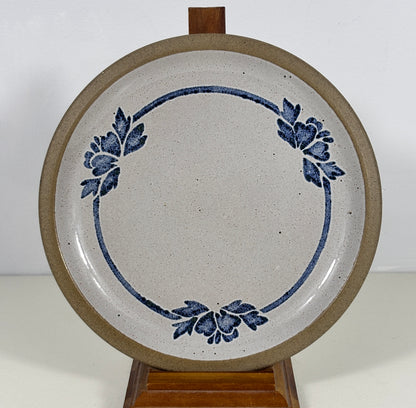 A round ceramic plate with a speckled off-white glaze, featuring a blue floral wreath design and a thick brown rim, displayed on a wooden stand against a plain white background.