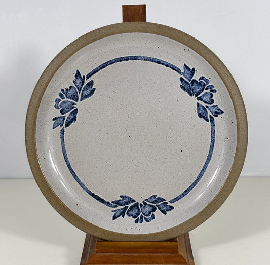 A round ceramic plate with a speckled off-white glaze, featuring a blue floral wreath design and a thick brown rim, displayed on a wooden stand against a plain white background.