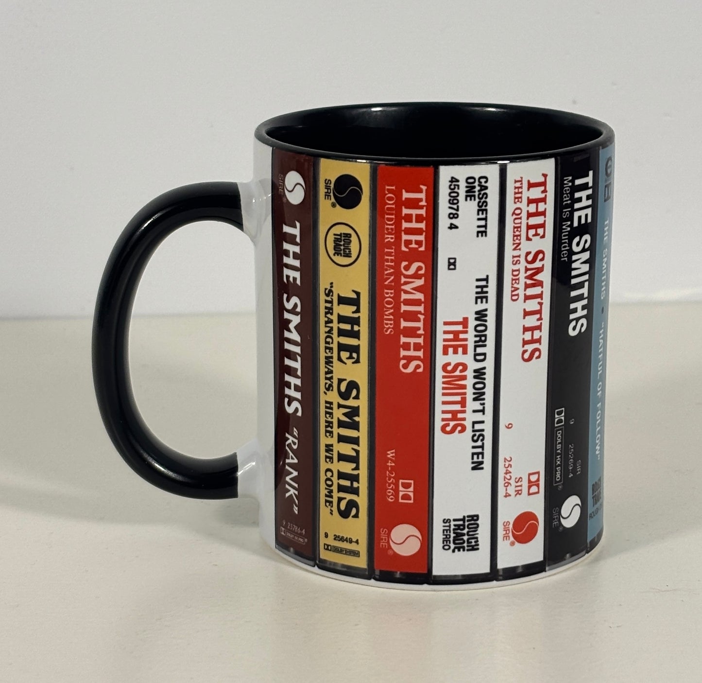 A ceramic mug featuring a printed design of various 'The Smiths' cassette tapes, with a solid black handle and interior, set against a plain light background.
