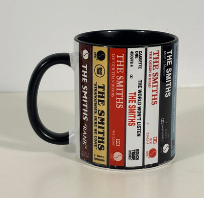A ceramic mug featuring a printed design of various 'The Smiths' cassette tapes, with a solid black handle and interior, set against a plain light background.
