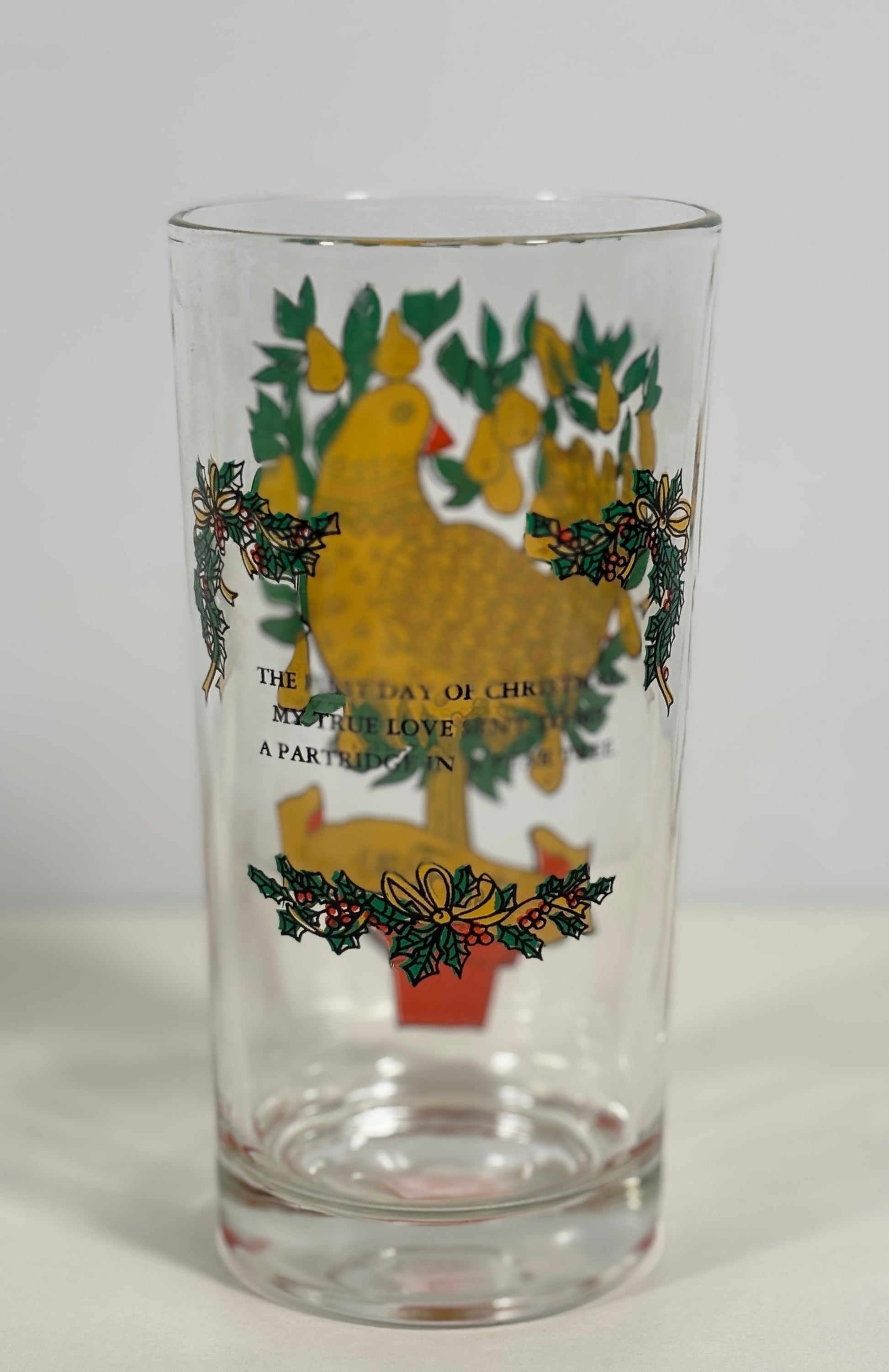 A clear glass drinking tumbler featuring a vintage-style Christmas design with a yellow turkey, green holly leaves, pinecones, and gold text on the front, set against a plain light gray background.