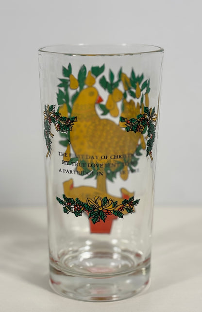 A clear glass drinking tumbler featuring a vintage-style Christmas design with a yellow turkey, green holly leaves, pinecones, and gold text on the front, set against a plain light gray background.