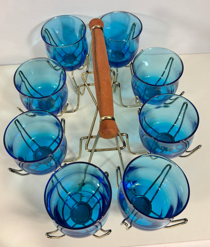 Eight blue tinted glass cups arranged around a metal stand with a wooden-handled tool, photographed on a plain white surface.