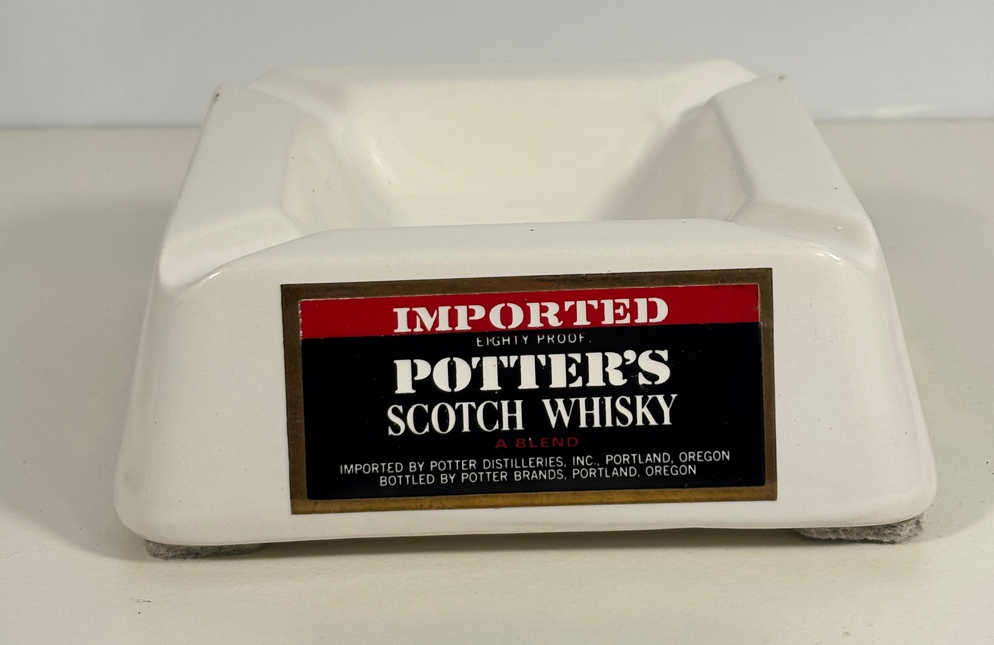 A white ceramic or porcelain potter's scotch whisky ashtray with a vintage-style label on the front reading 'IMPORTED POTTER'S SCOTCH WHISKY'.