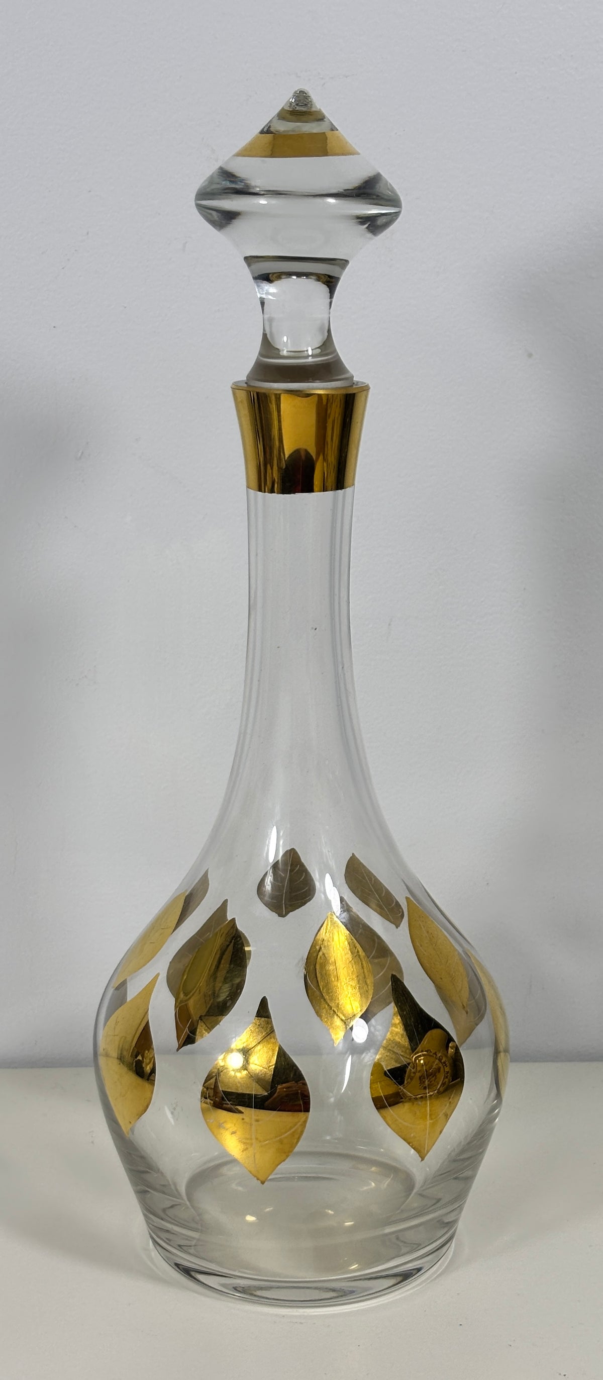 A clear glass decanter with a matching glass stopper, decorated with metallic gold leaf patterns, set against a plain light gray background.