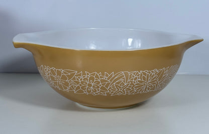 A vintage Pyrex mixing bowl in a warm tan color with a white floral pattern around the lower half and a white interior, shown against a neutral light gray background.