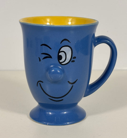 A blue ceramic mug with a winking cartoon face and a yellow interior, shown on a plain light background.