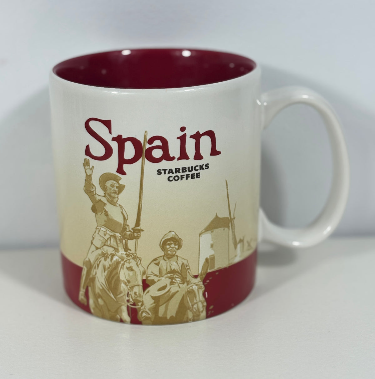 A white ceramic Starbucks Coffee mug with a dark red interior and bottom band, featuring a sepia-toned illustration of Spanish figures and the word 'Spain' on the front.