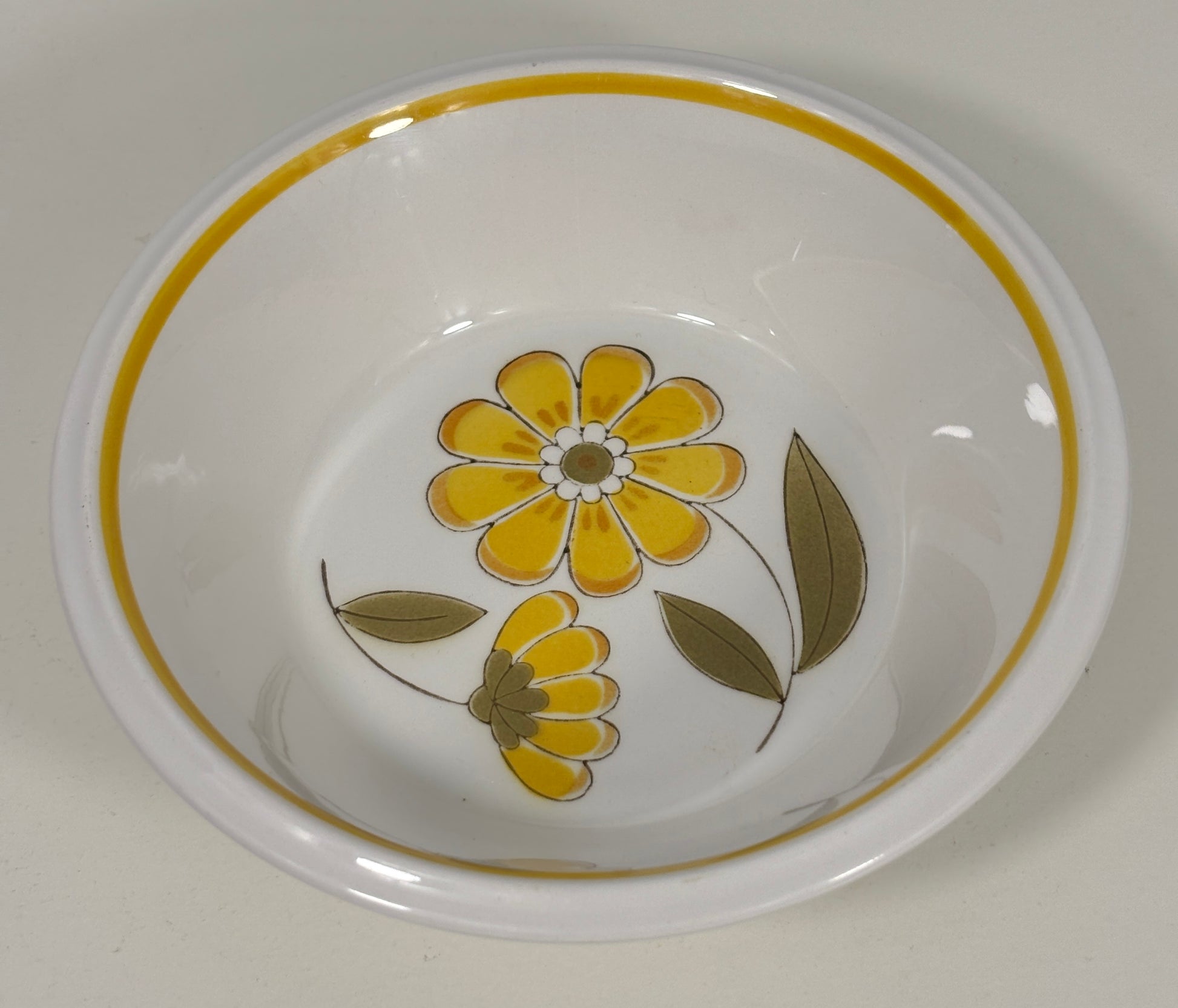 A white ceramic bowl featuring a yellow floral pattern and a yellow-gold rim, photographed from a top-down angle on a light neutral background.