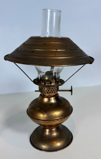 An antique brass oil lamp with a clear glass chimney and wick, set against a plain light gray background.
