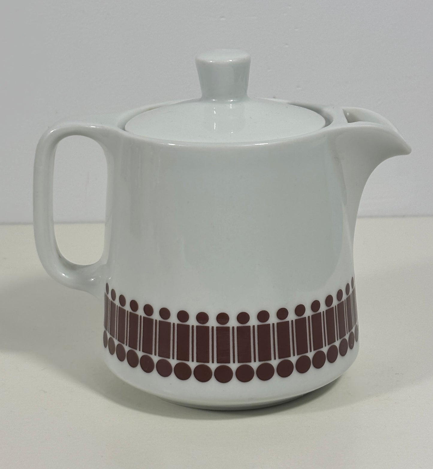 A white ceramic teapot with a lid, featuring a decorative band of dark brown vertical bars and dots near the base, set against a plain light background.