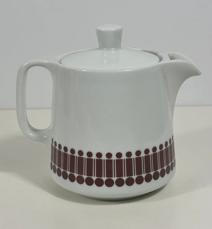 A white ceramic teapot with a lid, featuring a decorative band of dark brown vertical bars and dots near the base, set against a plain light background.
