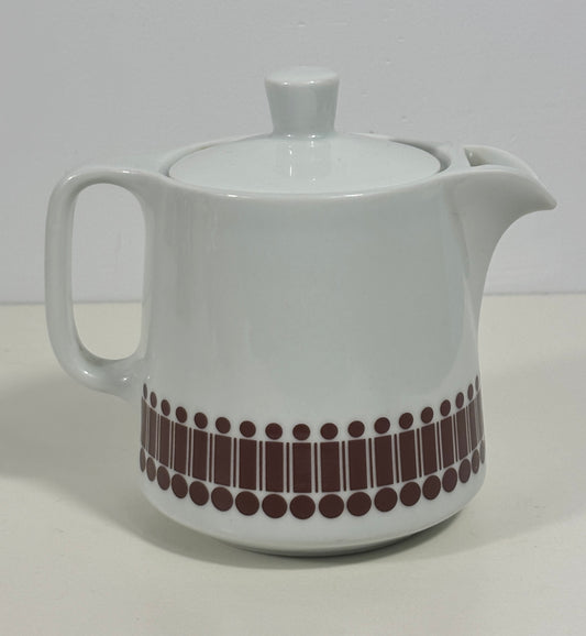 A white ceramic teapot with a lid, featuring a decorative band of dark brown vertical bars and dots near the base, set against a plain light background.