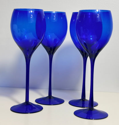 Four cobalt blue glass wine glasses with long stems and wide bowls, arranged on a white surface against a white background.