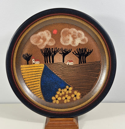 A round decorative ceramic plate with a hand-painted folk art landscape design, displayed on a wooden stand against a plain light gray background.