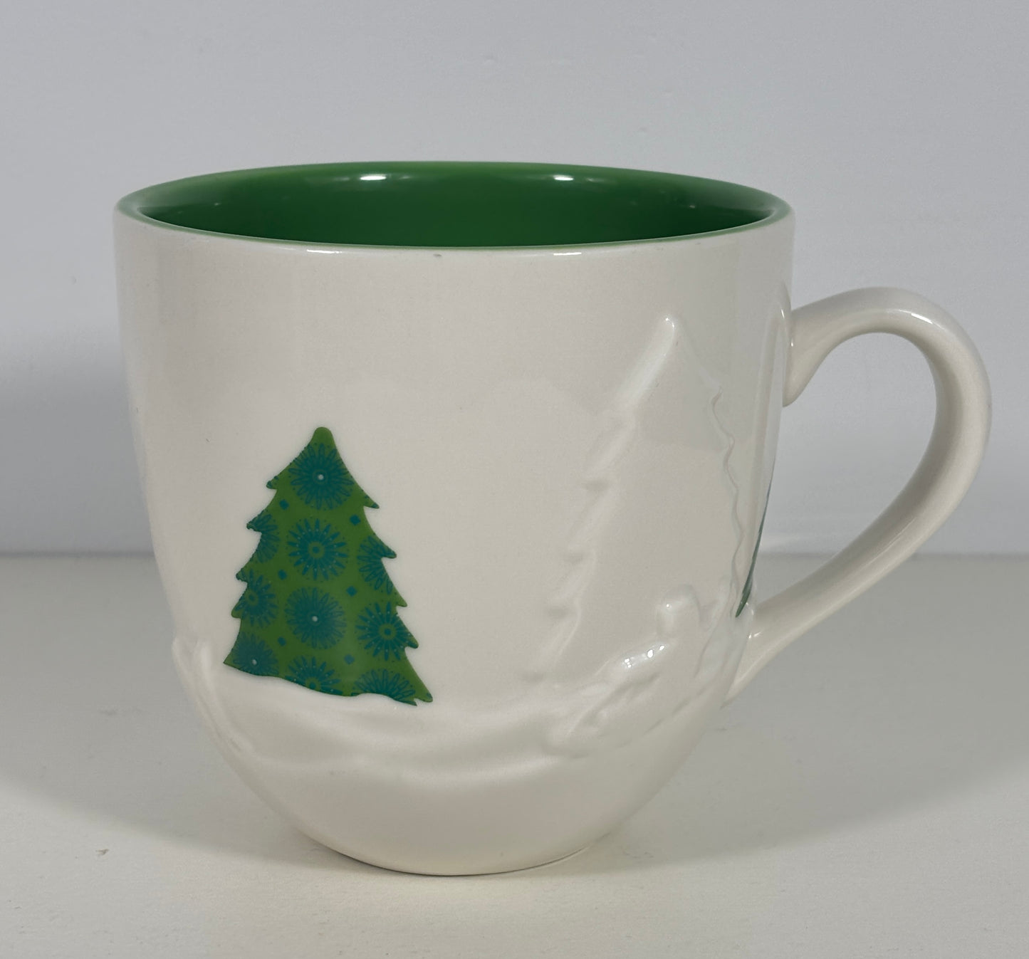 A white ceramic coffee mug with a dark green interior, featuring a green Christmas tree design and a white snowflake pattern on the exterior.