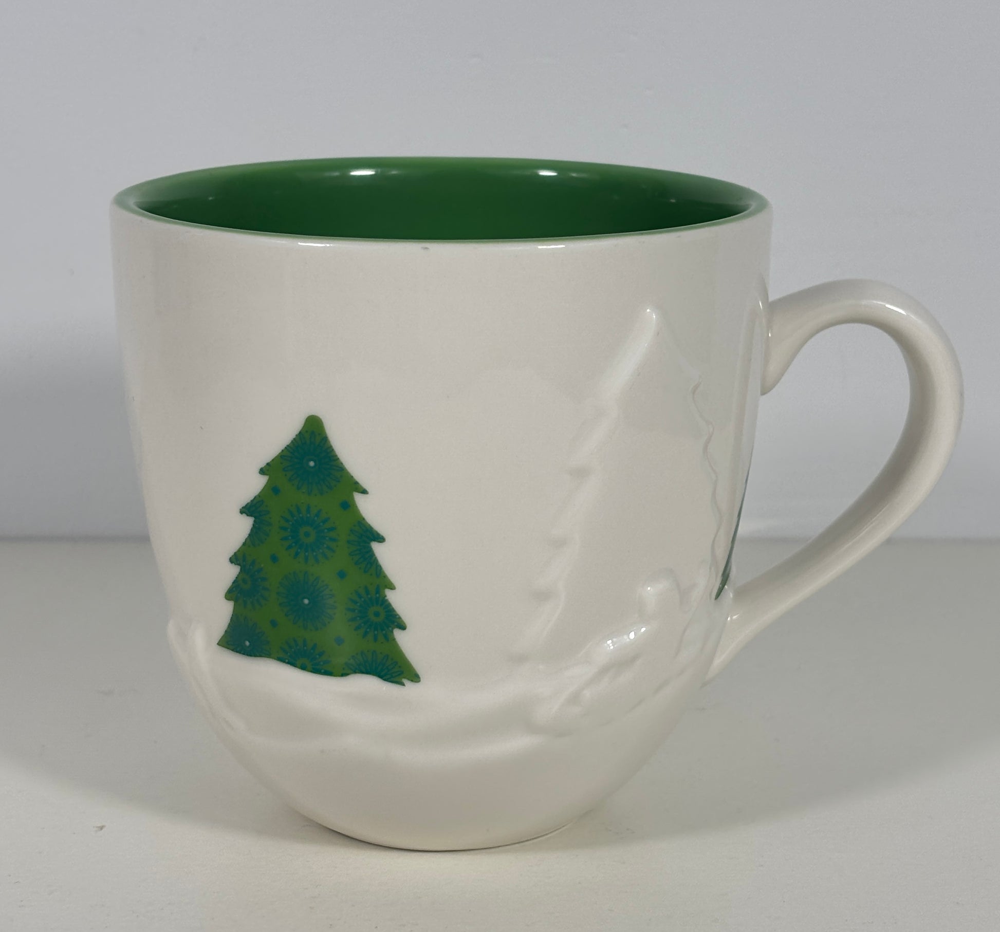 A white ceramic coffee mug with a dark green interior, featuring a green Christmas tree design and a white snowflake pattern on the exterior.