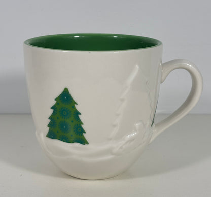 A white ceramic coffee mug with a dark green interior, featuring a green Christmas tree design and a white snowflake pattern on the exterior.