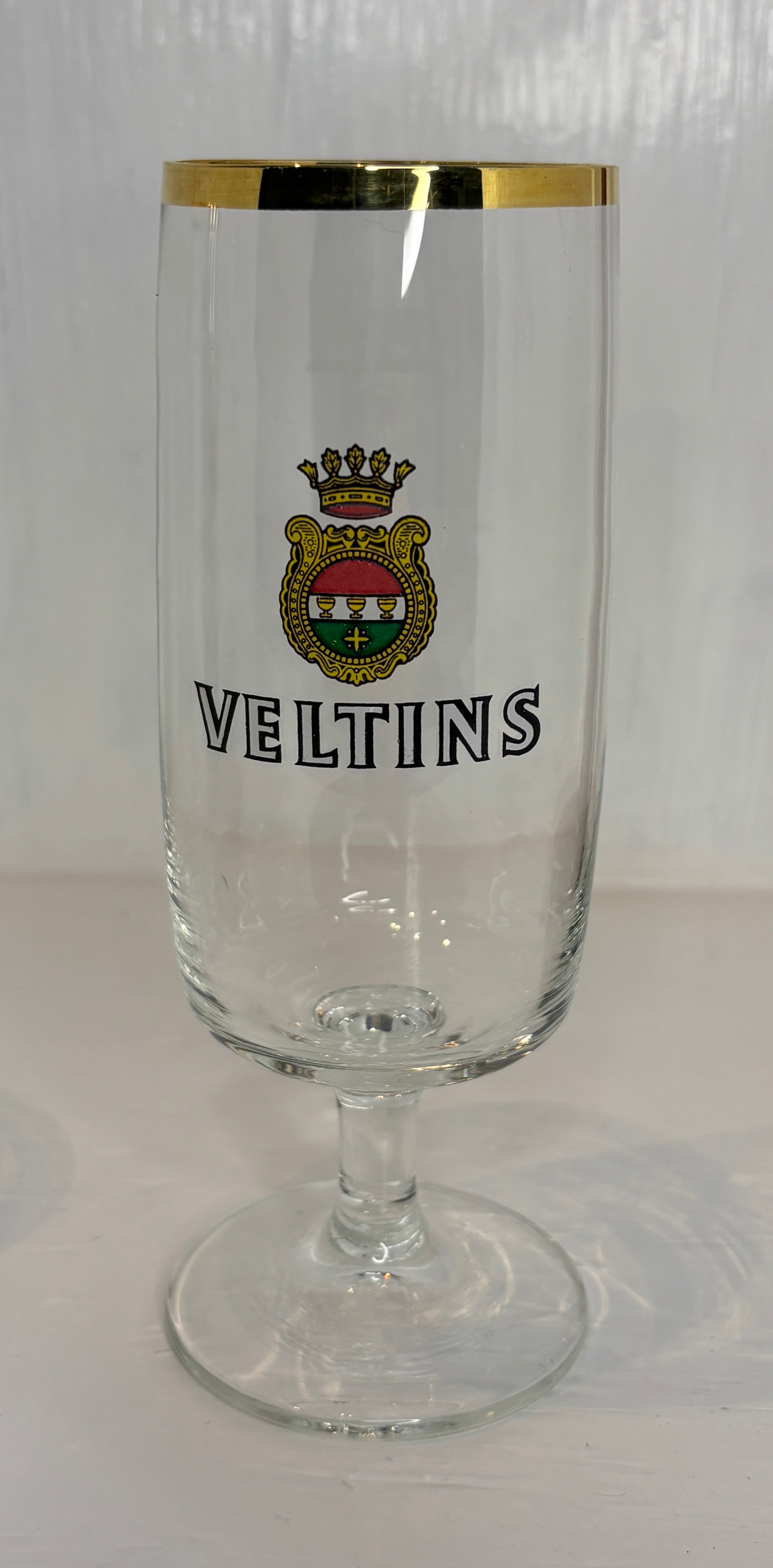 Clear glass beer mug with Veltins logo and gold rim on a white background