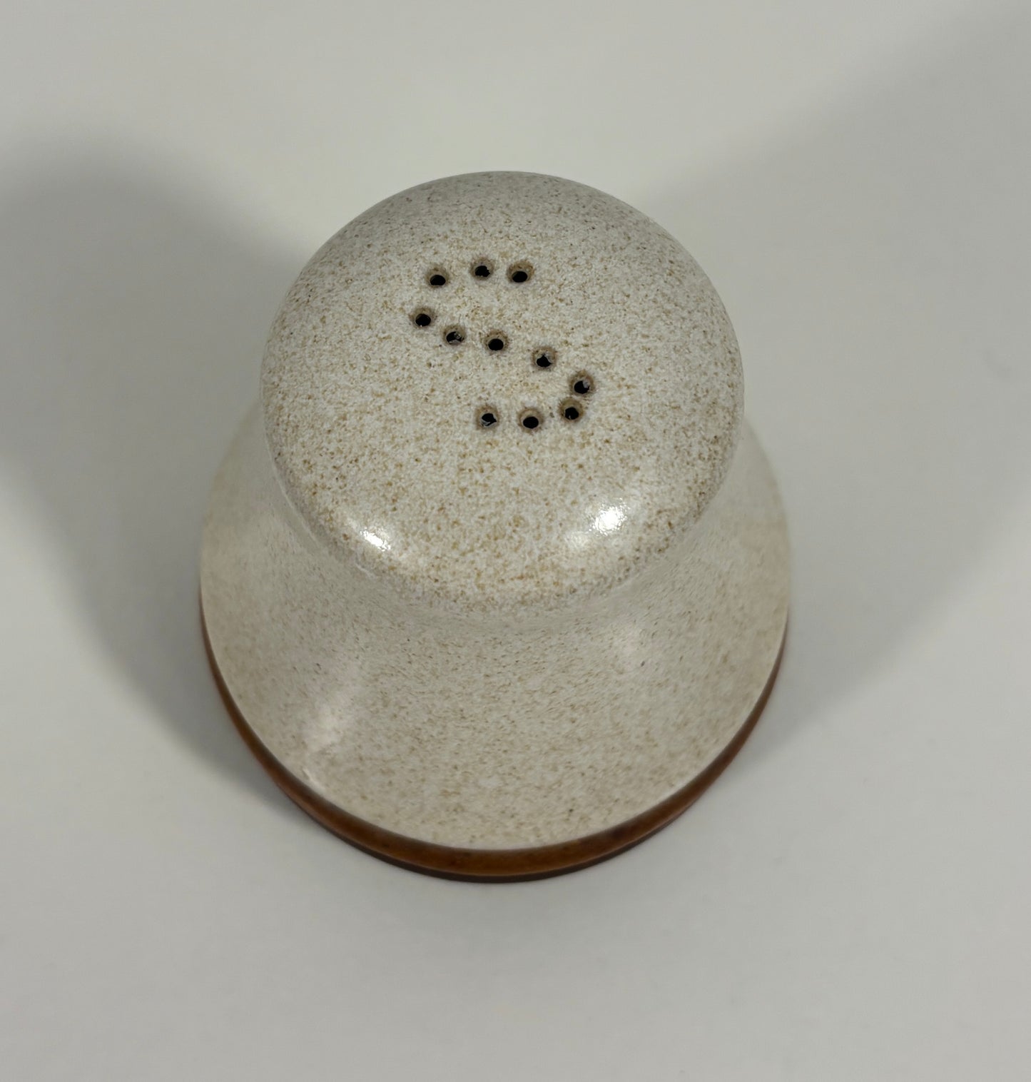 A ceramic salt or pepper shaker with a rounded, dome-shaped top featuring small perforated holes, finished in a speckled cream and brown glaze with a solid brown base, shown on a neutral light gray background.