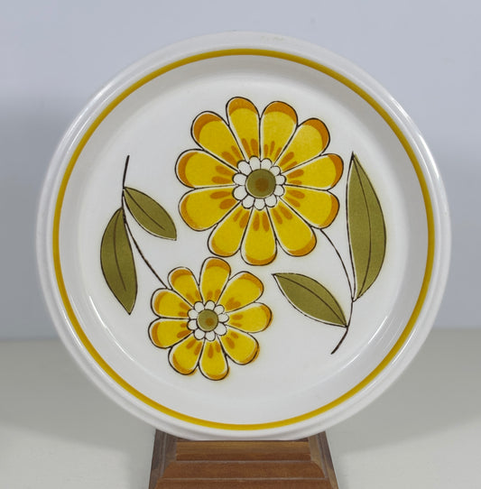 A round white ceramic plate with a retro floral pattern featuring yellow flowers and green leaves, displayed on a wooden stand against a light gray background.