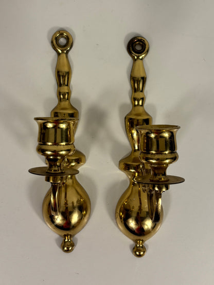 A pair of polished brass wall-mounted candle holders with decorative scrollwork and looped top and bottom details, shown against a plain off-white background.