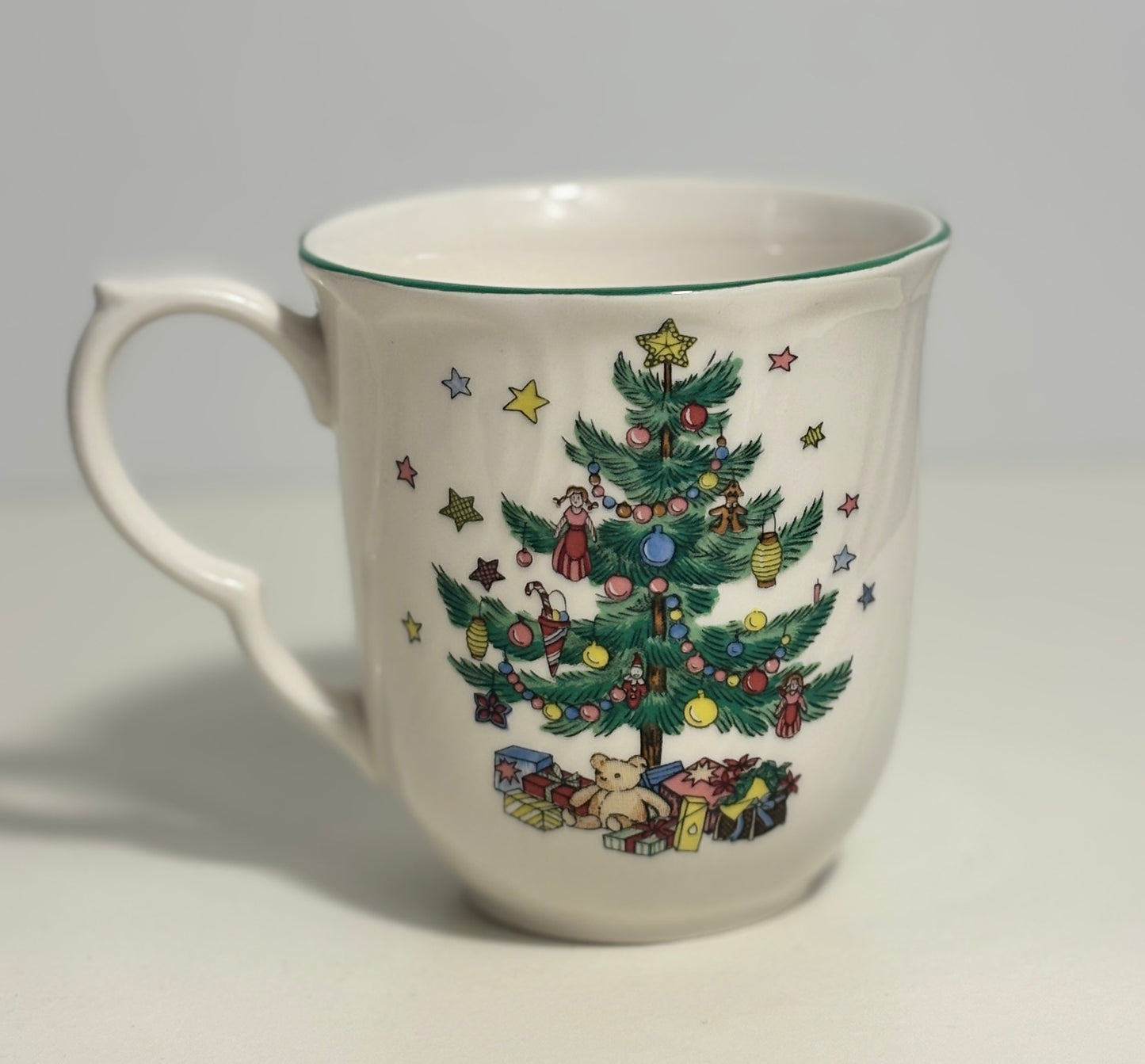 A white ceramic mug featuring a colorful Christmas tree illustration decorated with ornaments, stars, gifts, and small figurines, set against a plain light gray background.