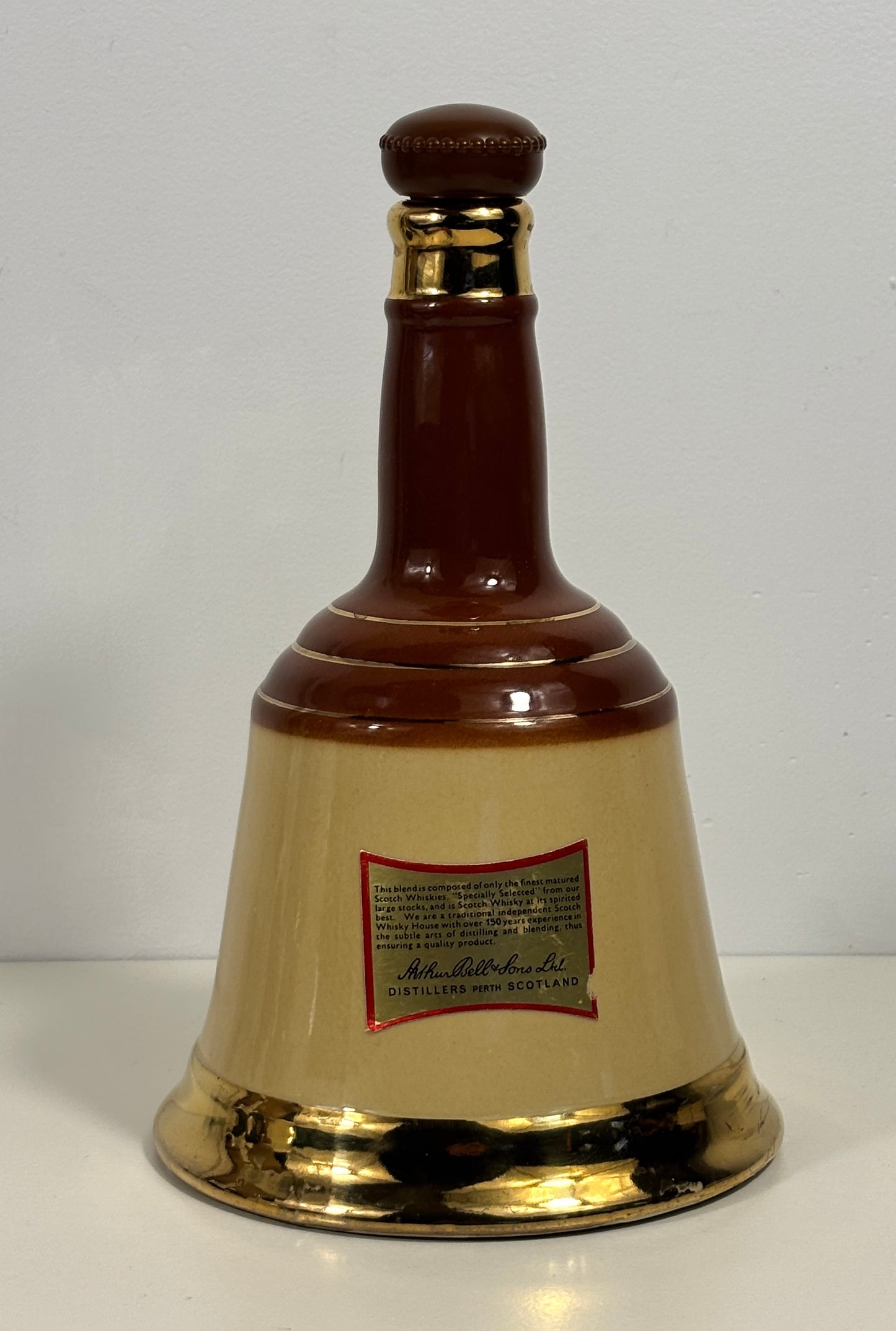 A decorative ceramic bell-shaped bottle with a glossy dark brown neck and base, a cream-colored center body, and metallic gold accents at the neck collar and bottom rim. A small red and gold label is attached to the front.