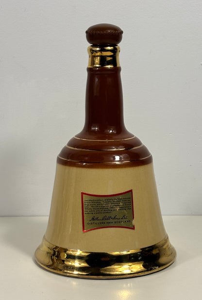 A decorative ceramic bell-shaped bottle with a glossy dark brown neck and base, a cream-colored center body, and metallic gold accents at the neck collar and bottom rim. A small red and gold label is attached to the front.