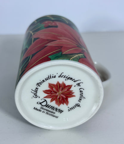 A close-up of a Duneon 'Golda Painsellia' designed by Caroline Dressler stoneware mug, showing the white printed base with a red poinsettia star design and text, with the rest of the festive red and green poinsettia pattern visible on the side.