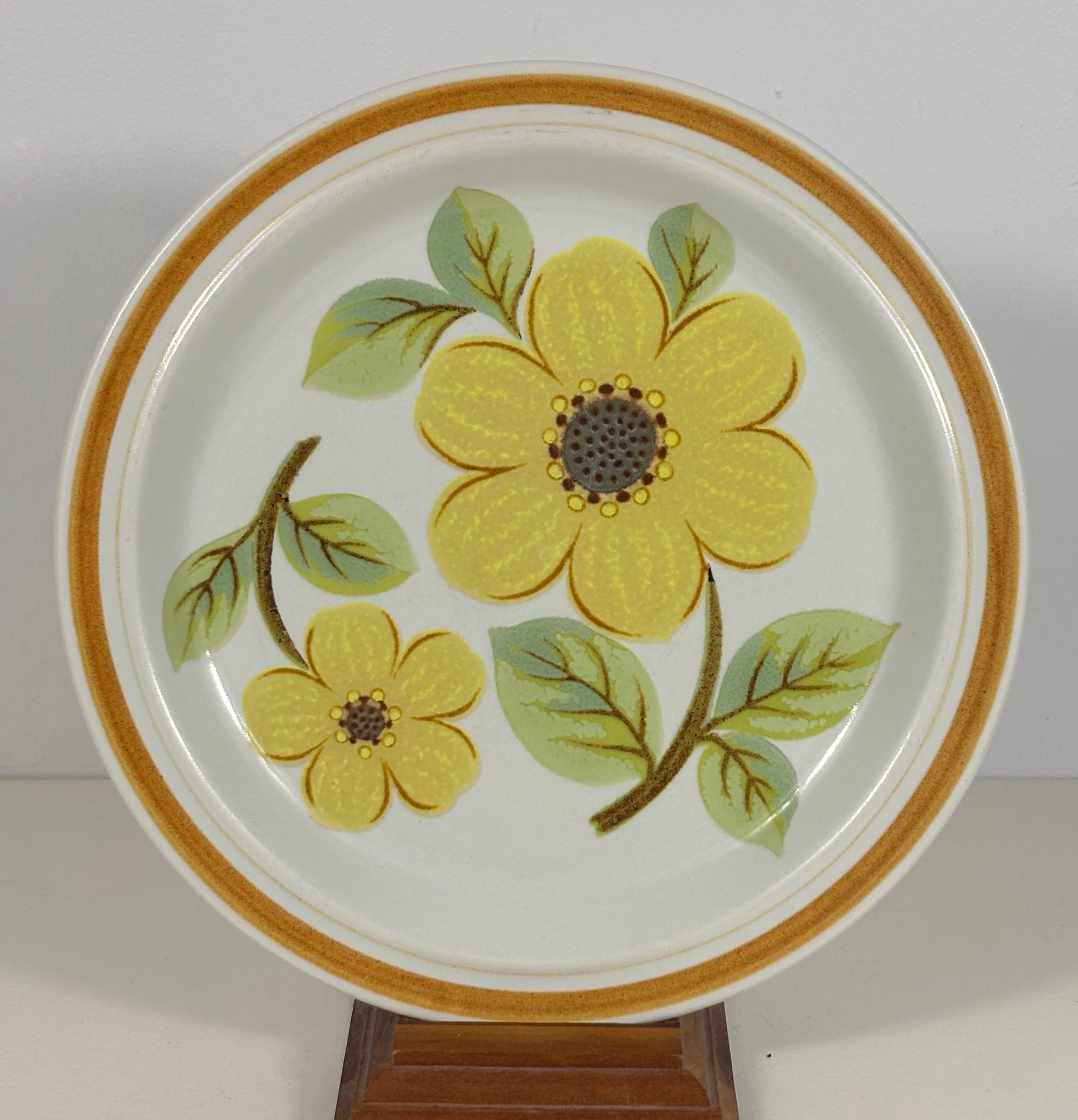 A round white ceramic plate featuring a hand-painted yellow flower and green leaves, with a gold-toned rim, displayed on a wooden stand against a plain light background.