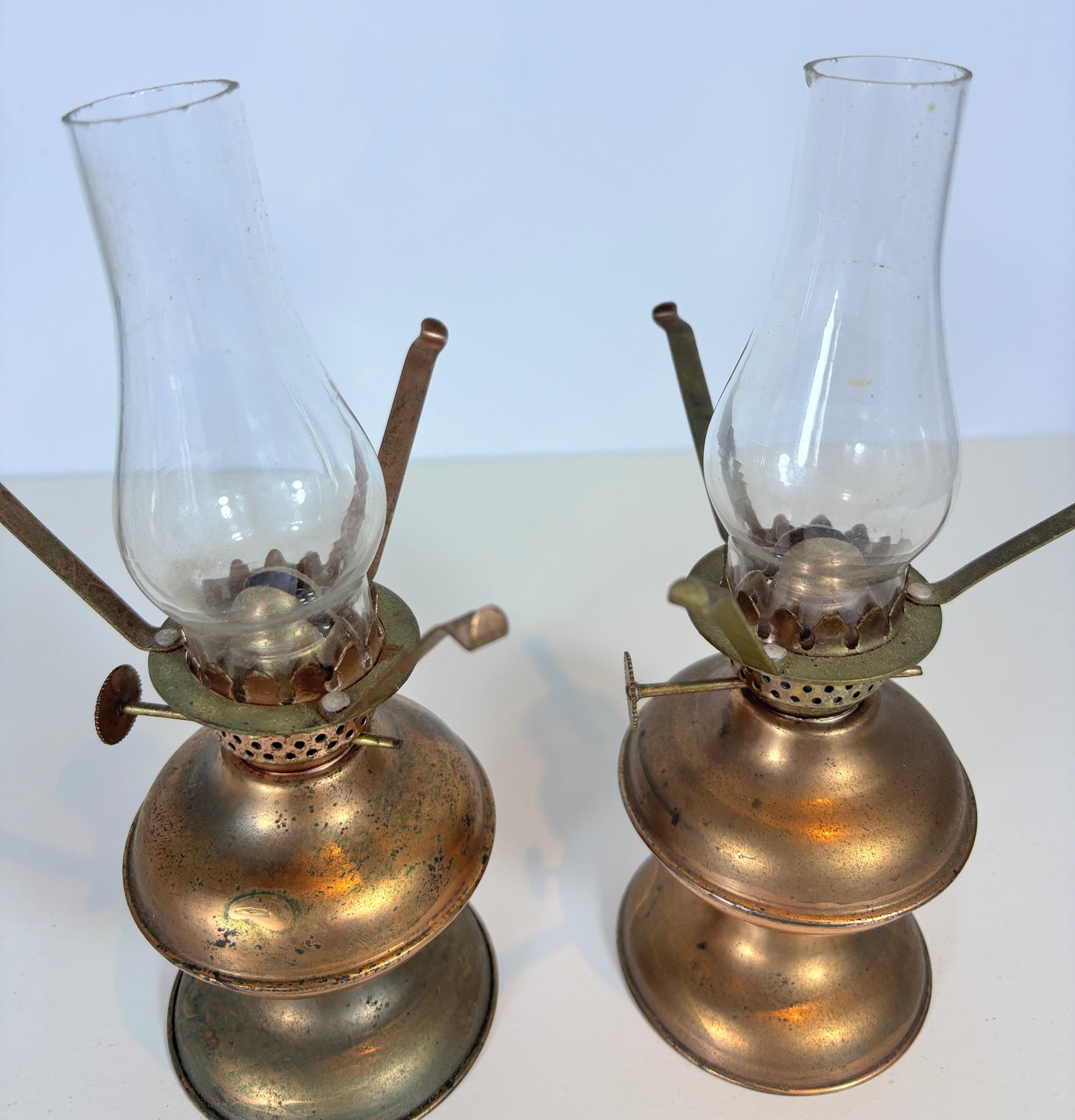 Two vintage brass oil lamps with clear glass burners, displayed side by side on a white surface against a light background.