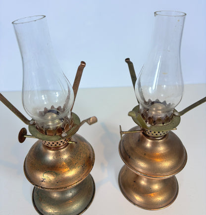 Two vintage brass oil lamps with clear glass burners, displayed side by side on a white surface against a light background.