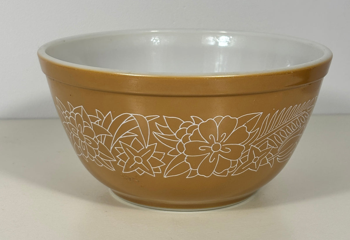 A vintage Pyrex mixing bowl in a warm tan-brown color with a white floral line-art pattern on the outside and a white glazed interior.