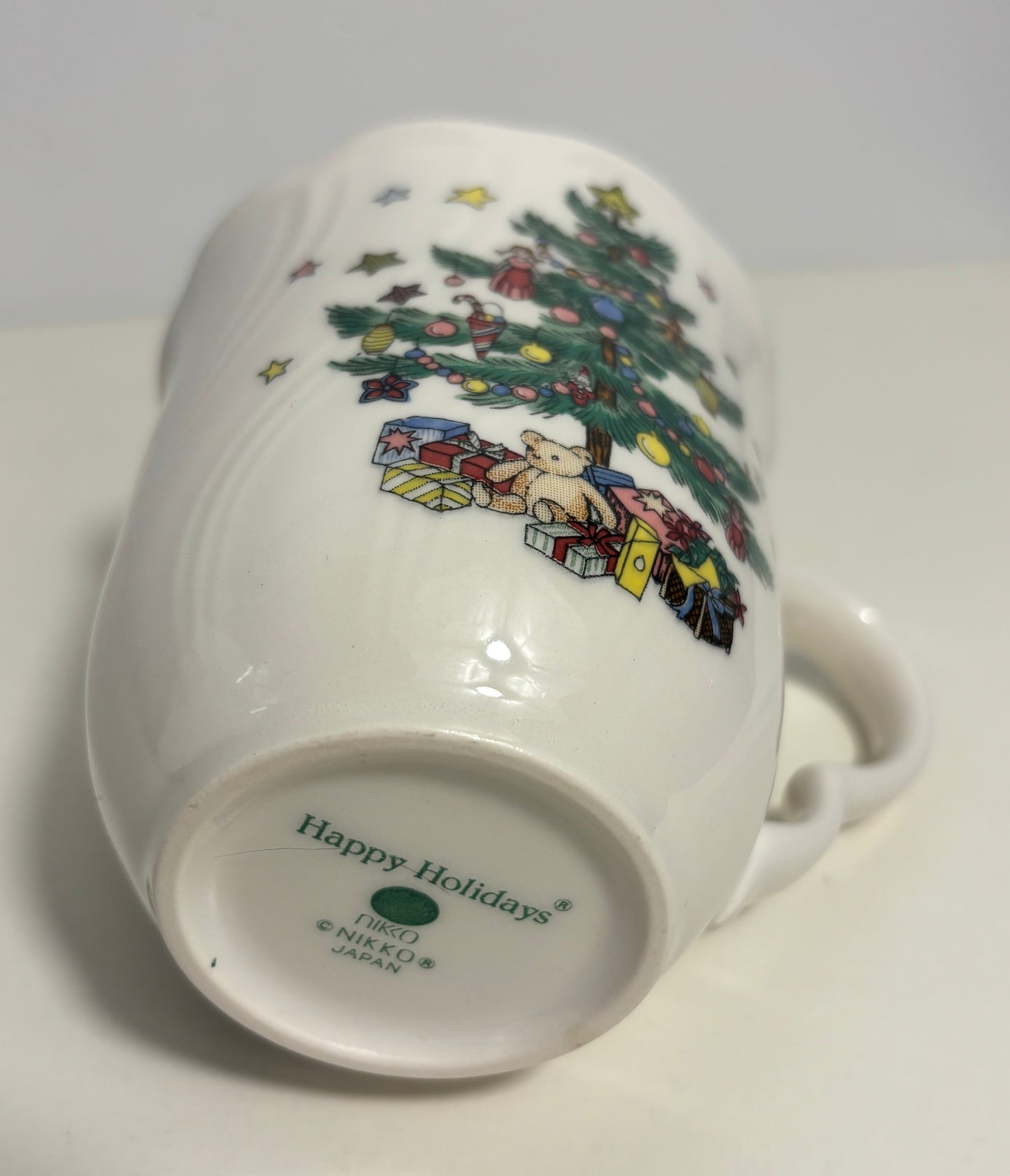 A white ceramic mug shown from its bottom side, featuring a colorful illustration of a decorated Christmas tree and wrapped gifts. The inside bottom is printed with 'Happy Holidays' and 'Nikko Japan'.