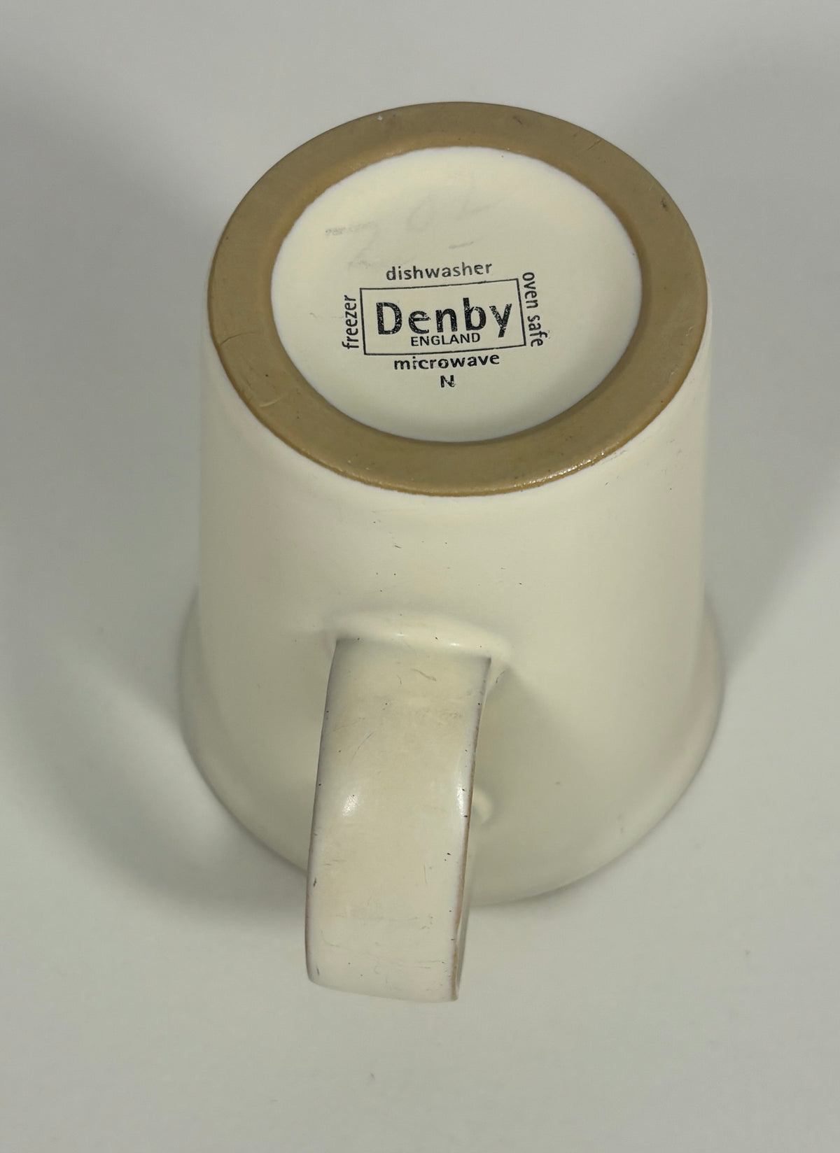 An off-white ceramic mug shown from its bottom side, featuring a gold-colored rim and a handle. The inside bottom is printed with black text including 'Denby' and 'dishwasher'.