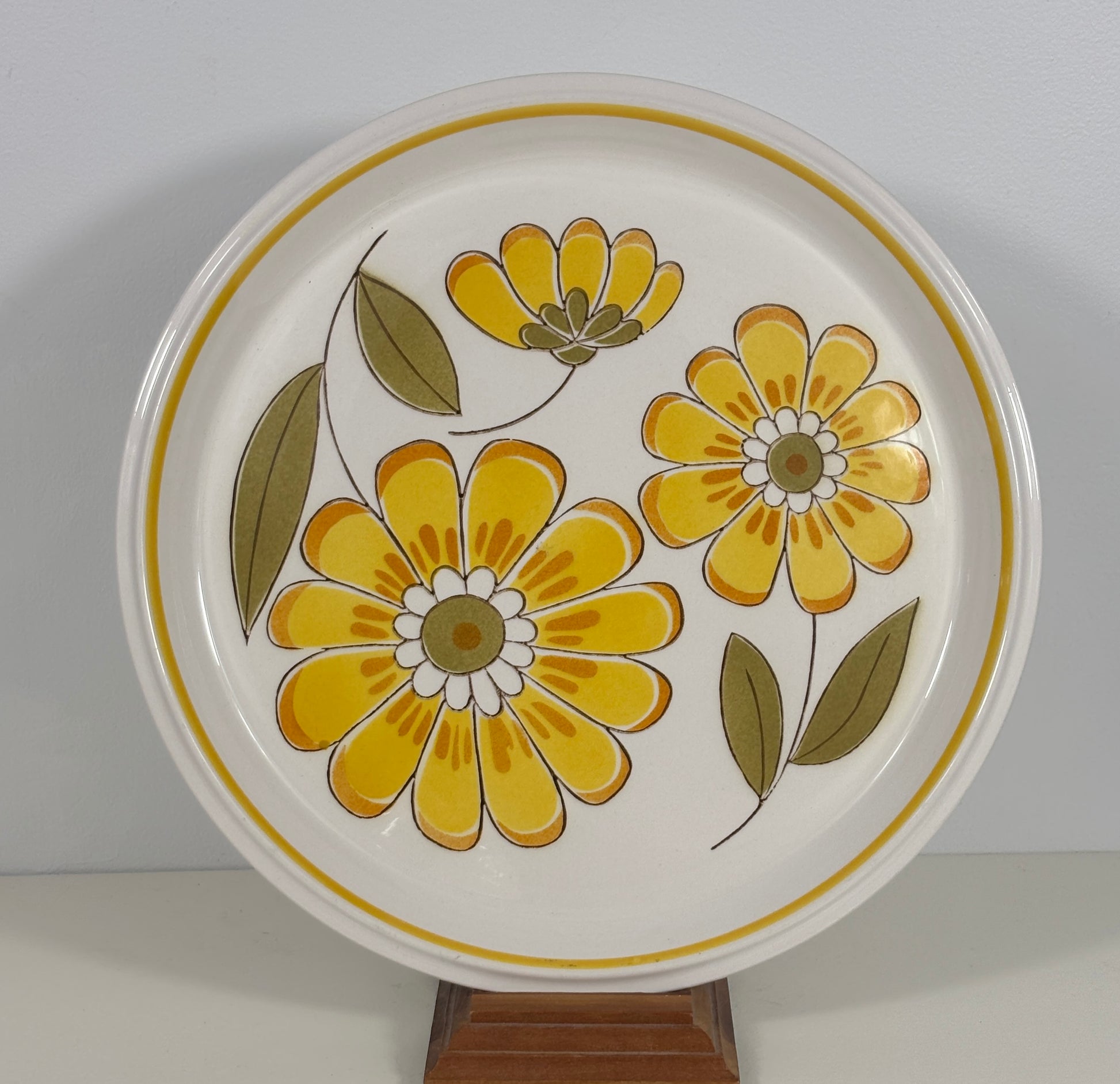 A round white ceramic plate with a retro floral pattern featuring yellow flowers and olive green leaves, displayed on a wooden stand against a neutral gray background.