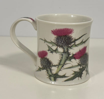 A white ceramic mug featuring a detailed botanical print of thistles with pink-purple flowers and green leaves, set against a plain light gray background.