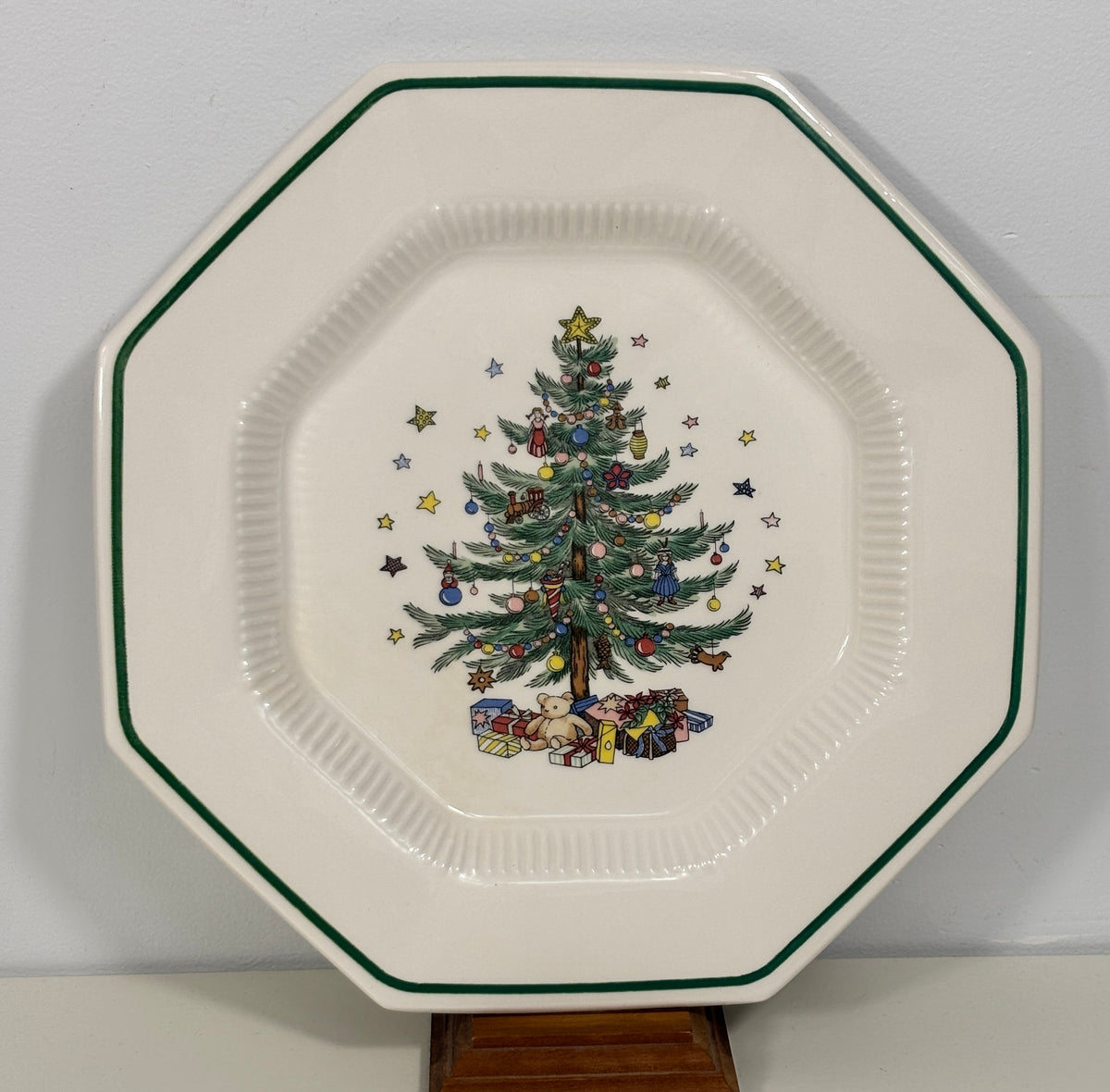 An octagonal white ceramic plate with a green rim, featuring a colorful Christmas tree illustration in the center, displayed on a wooden stand against a light gray wall.
