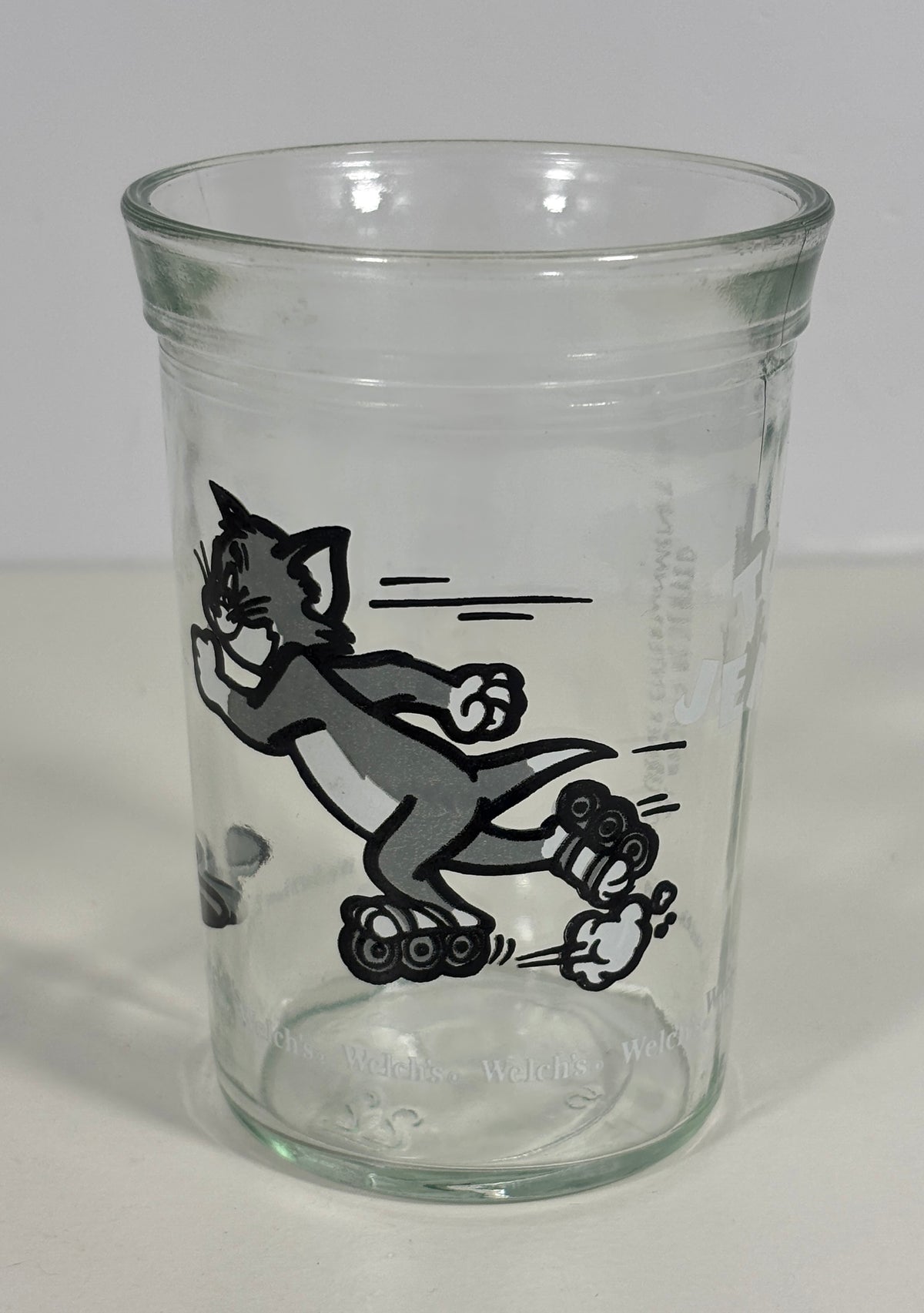 A clear glass tumbler featuring a black-and-white printed illustration of Tom and Jerry cartoon characters, photographed against a plain light background.