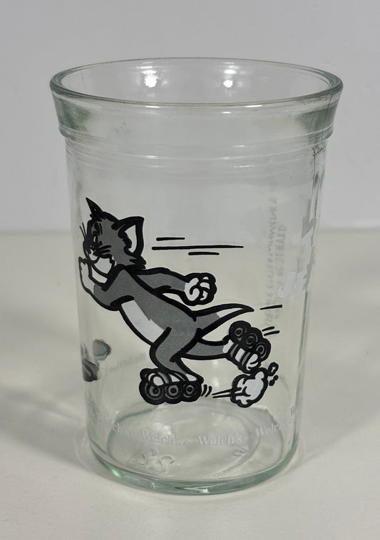 A clear glass tumbler featuring a black-and-white printed illustration of Tom and Jerry cartoon characters, photographed against a plain light background.